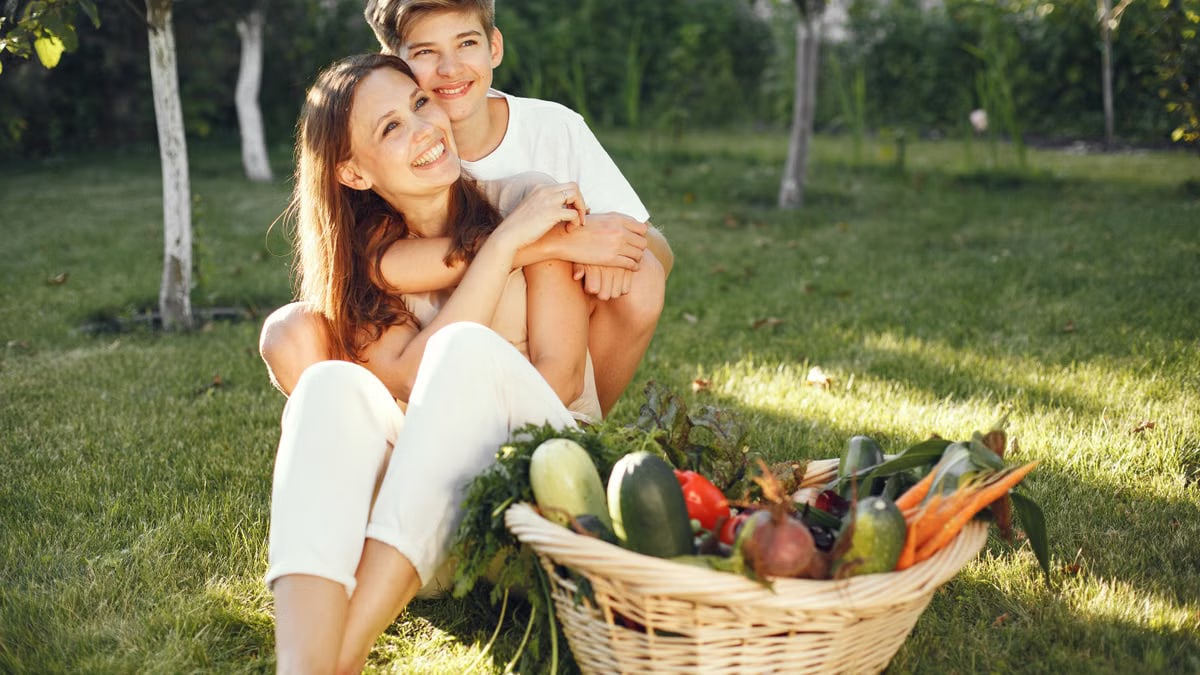 A joyful mother and son sitting on grass with a basket of fresh vegetables.